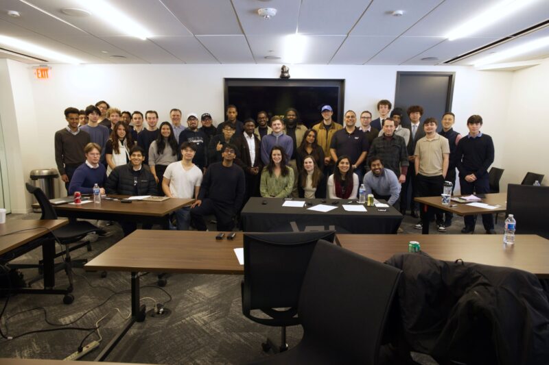 A diverse group of people stand and sit in a meeting room.