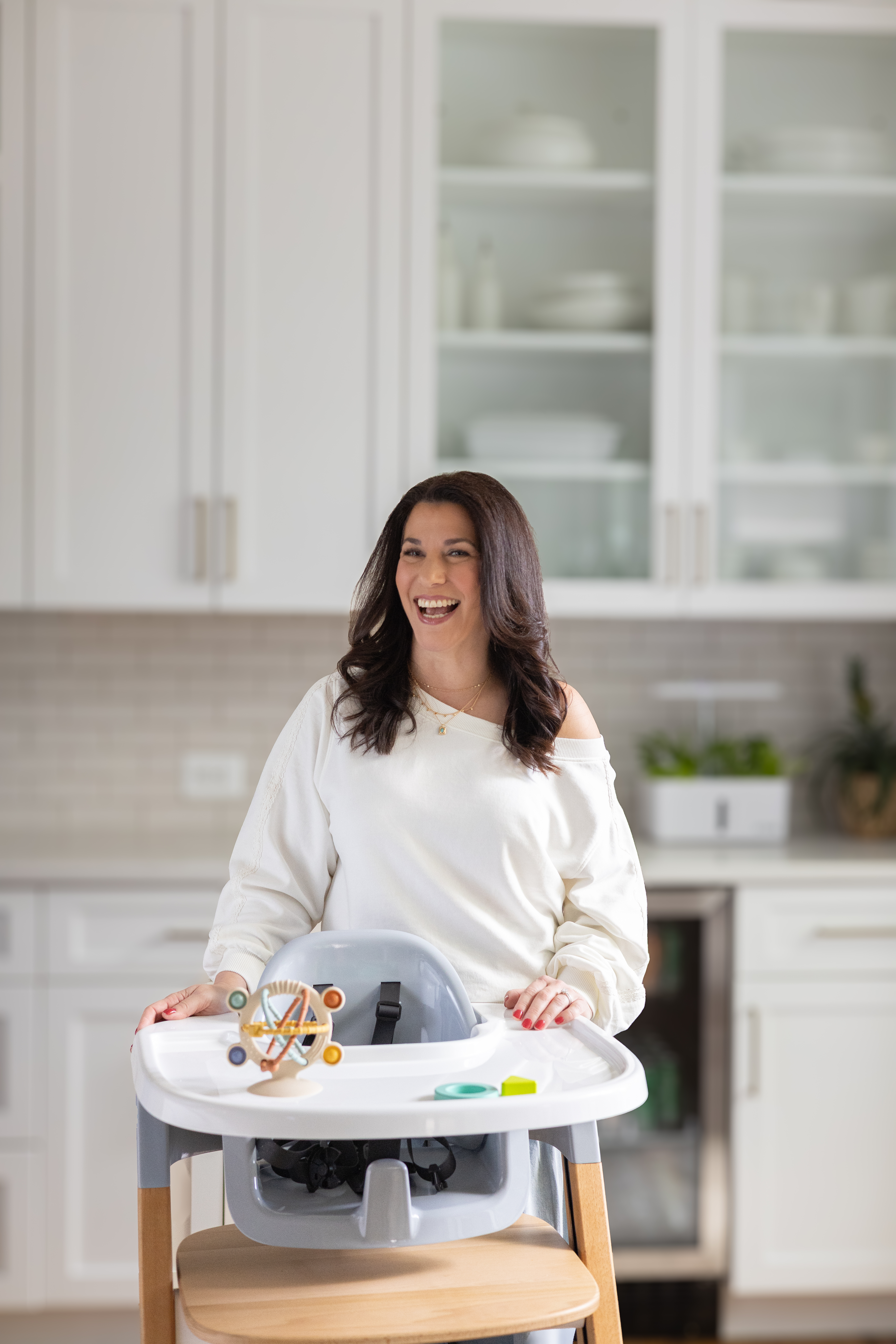 A woman in a white sweater stands and smiles behind a baby high chair in a modern kitchen with white cabinets and dishes.