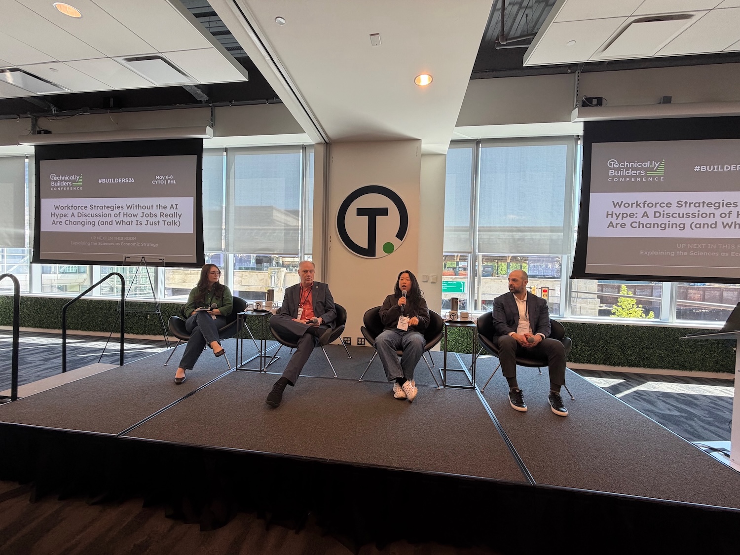 Four panelists sit on stage in front of an audience at a conference. A screen behind them displays information about a discussion on workforce strategies and AI.