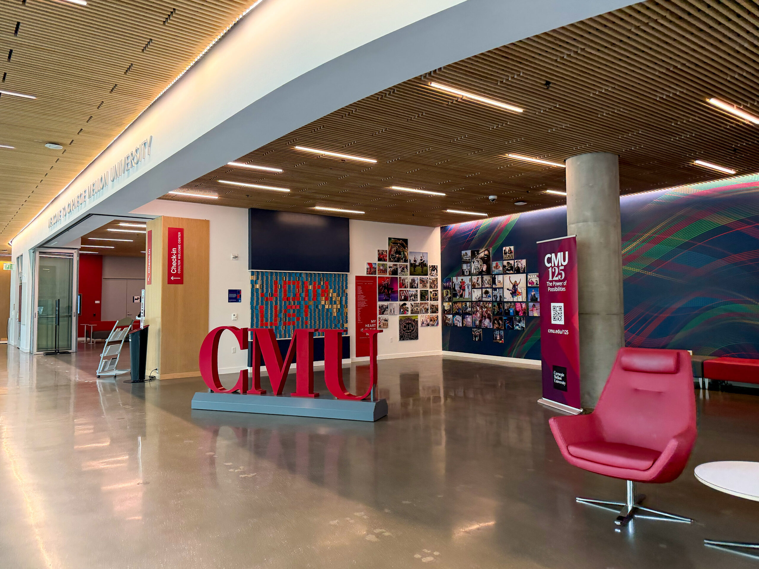 Modern university lobby with red CMU letters, wall art, photos, and signage, featuring red chairs and a wavy-patterned accent wall.