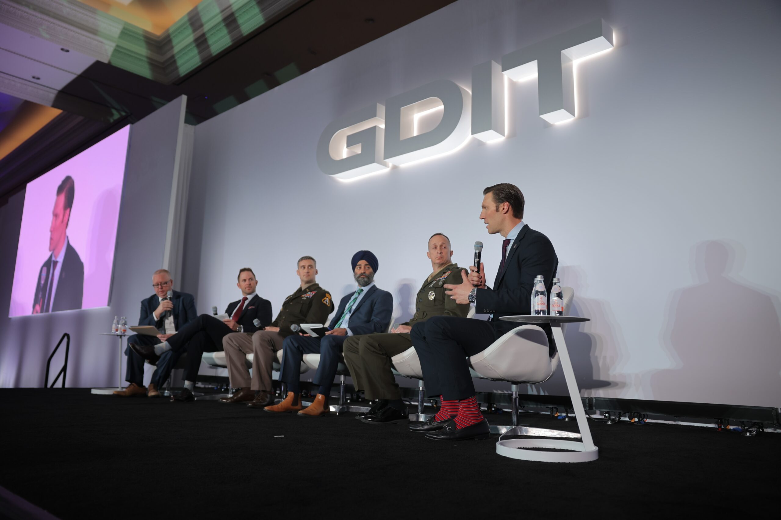 Six men sit on a stage panel at a GDIT event, with one man speaking into a microphone while others listen. A large GDIT logo is displayed behind them.