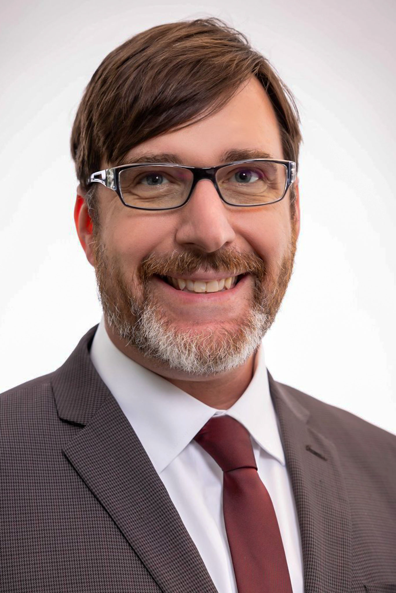 A man with glasses, brown hair, and a beard, wearing a brown suit, white shirt, and maroon tie, smiles at the camera against a plain light background.