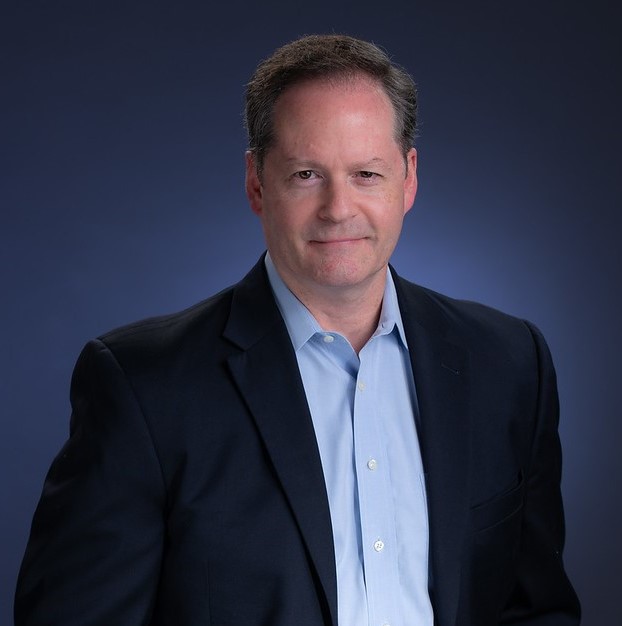 A middle-aged man wearing a dark suit jacket and light blue shirt poses in front of a dark blue background.