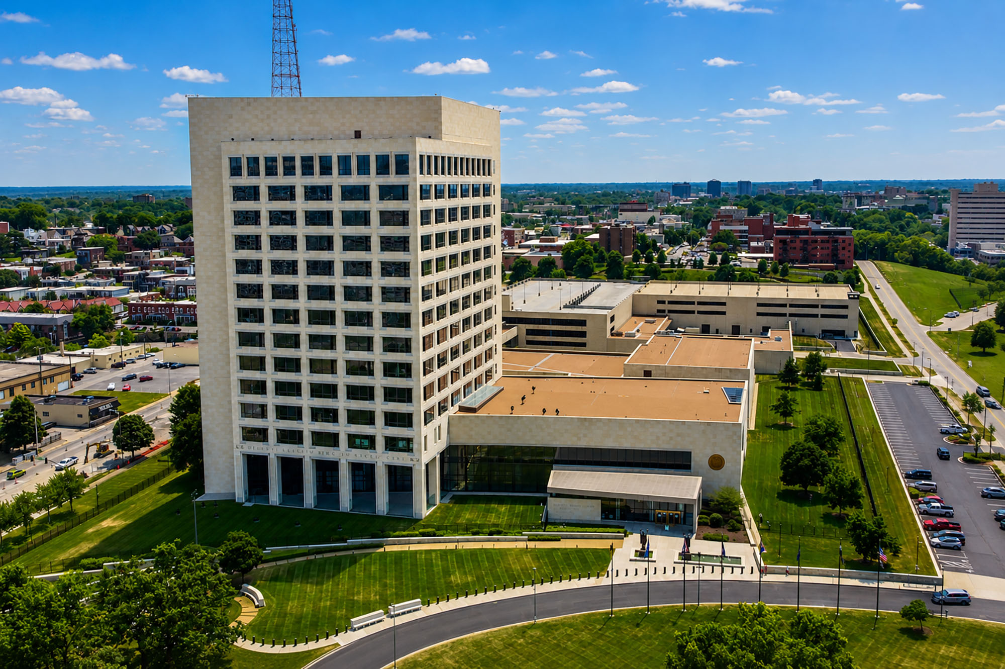 Aerial view of a tall, rectangular office building with adjacent structures, surrounded by green lawns, trees, and parking lots under a blue sky.