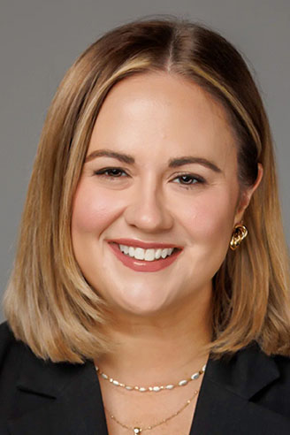 A woman with straight, shoulder-length blonde hair, wearing a black blazer and gold jewelry, smiles at the camera against a plain gray background.
