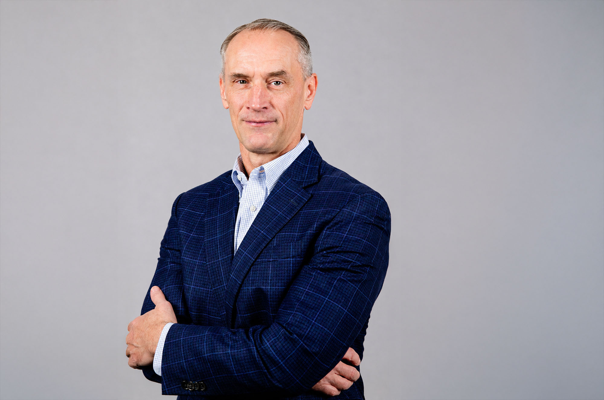 A middle-aged man with short gray hair wearing a blue plaid suit jacket and light shirt stands with arms crossed against a plain gray background.