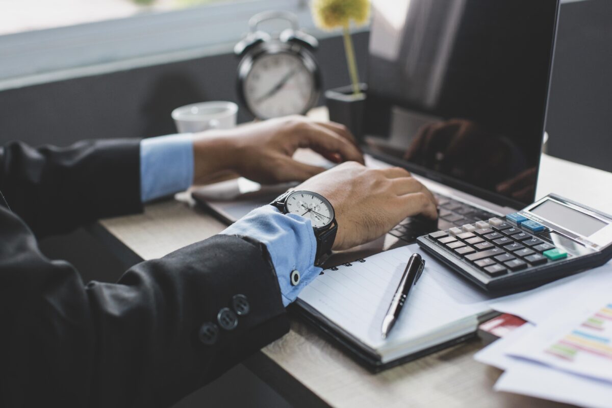 A person in a suit works on a laptop at a desk with a calculator, pen, notebook, papers, and an alarm clock.