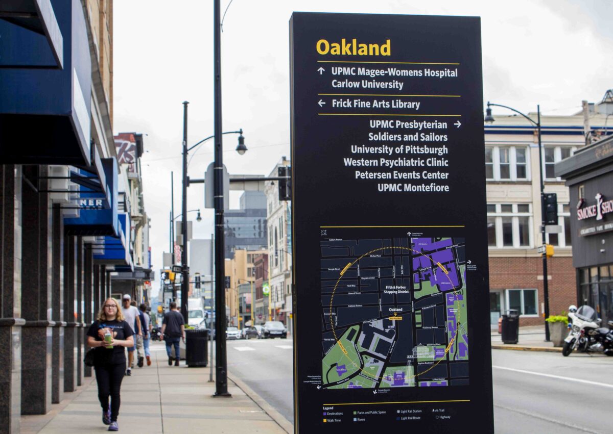 A directional sign in Oakland, Pittsburgh lists local institutions and shows a map; pedestrians walk along the street with shops and buildings in the background.
