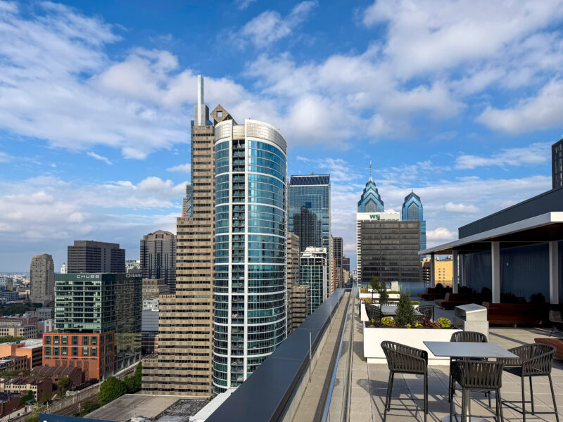 Rooftop terrace with tables and chairs overlooking a city skyline of modern high-rise buildings under a partly cloudy sky.