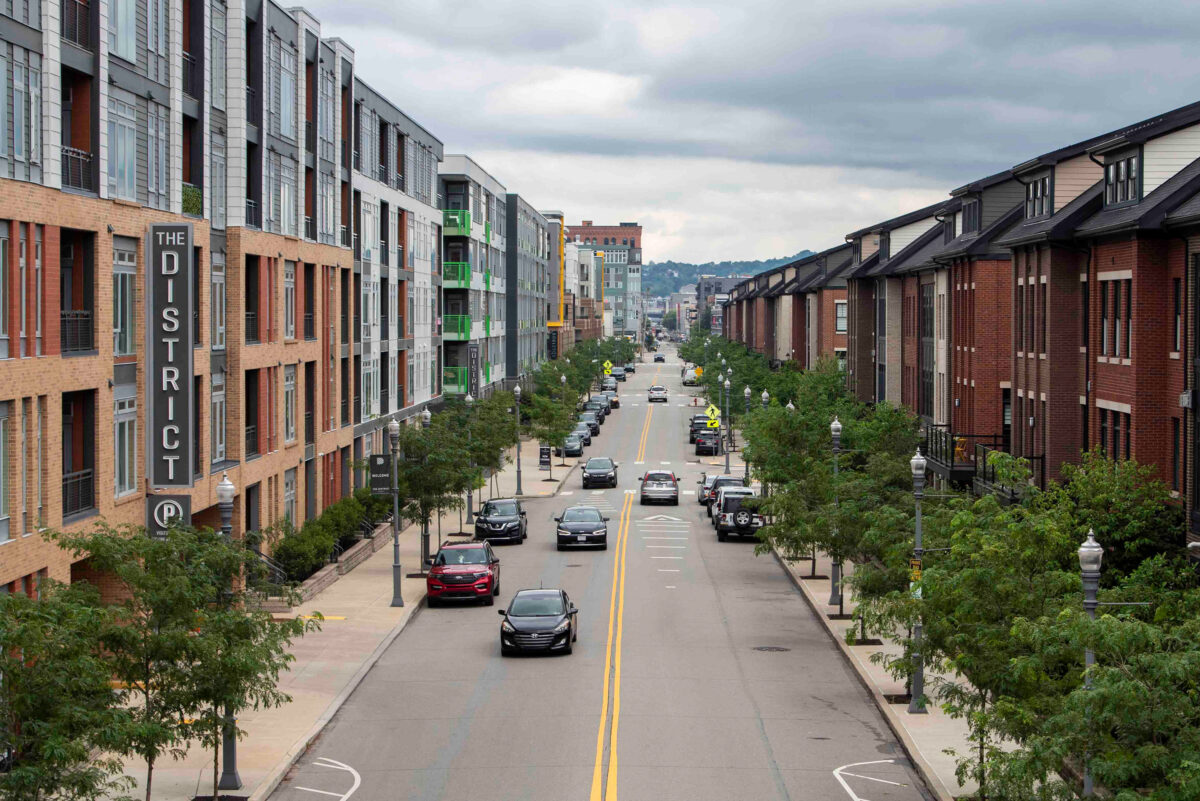 A city street lined with modern apartment buildings, parked cars, trees, and a sign reading "The District" on the left side under a cloudy sky.