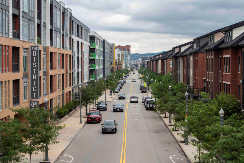 A city street lined with modern apartment buildings, parked cars, trees, and a sign reading "The District" on the left side under a cloudy sky.