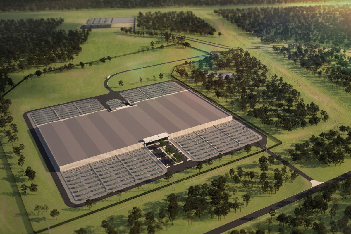 Aerial view of a large industrial warehouse or factory surrounded by parking lots and green wooded areas.