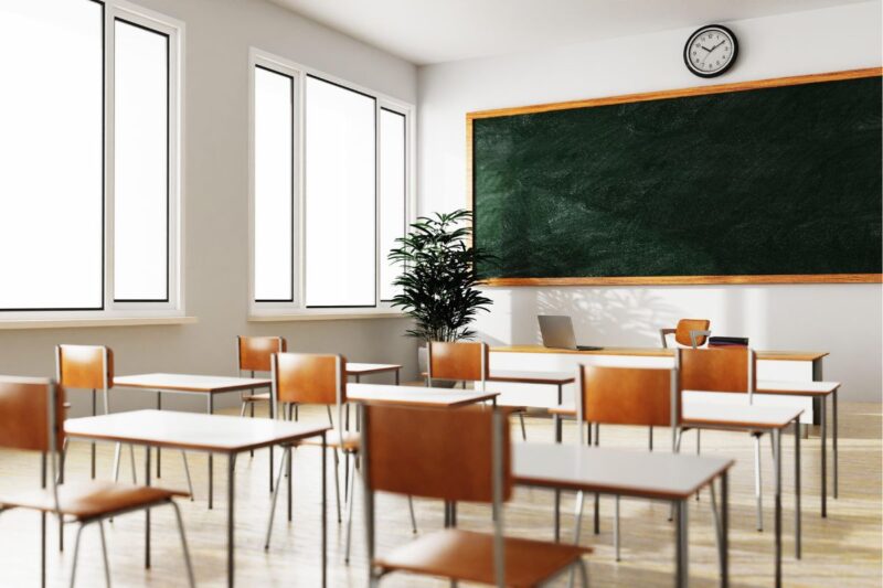 Empty classroom with wooden desks and chairs, a large green chalkboard, a clock on the wall, large windows, and a potted plant near the front.