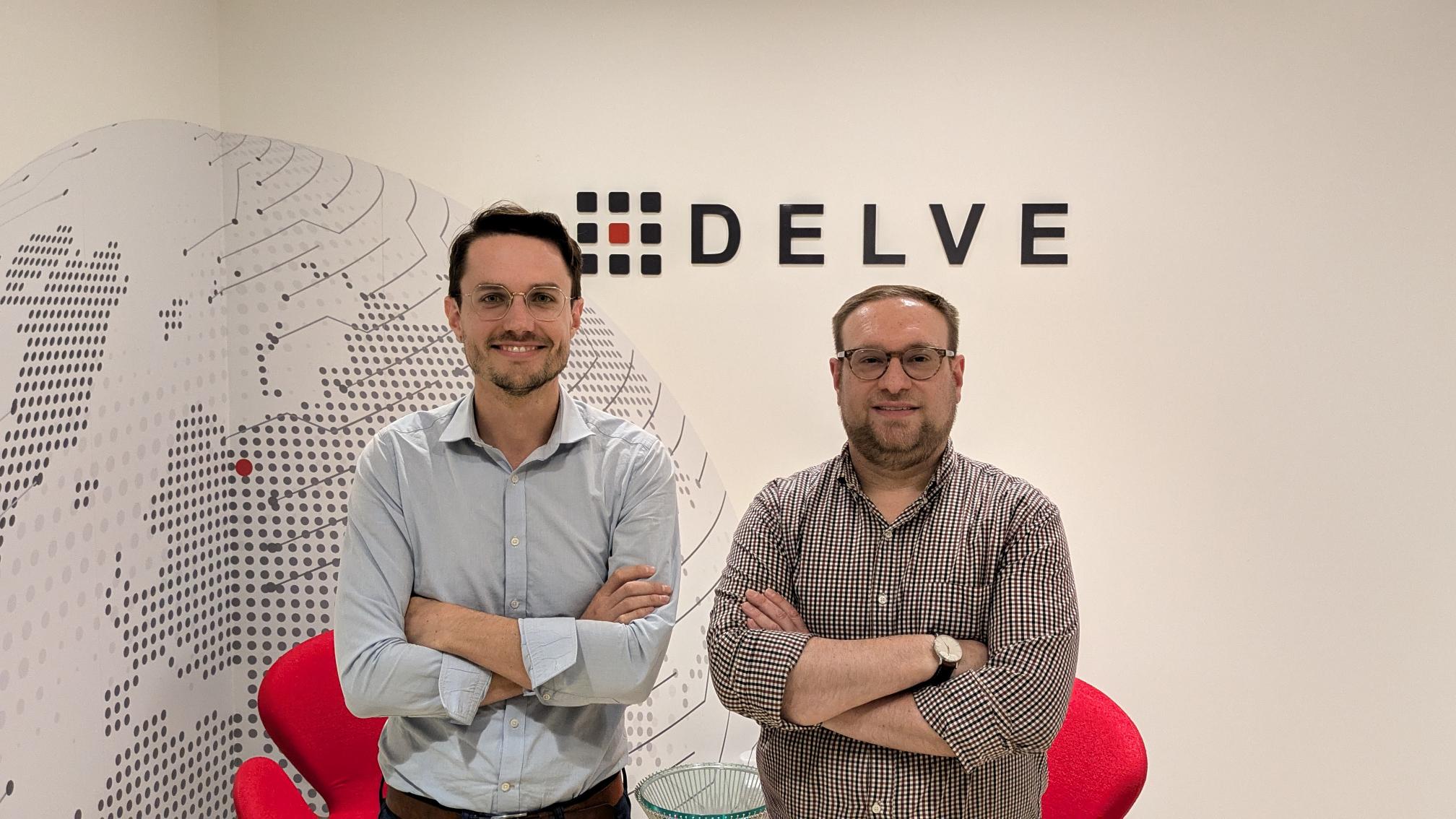 Two men stand with arms crossed in front of a wall featuring the DELVE logo and abstract design, with two red chairs and a glass table in the background.