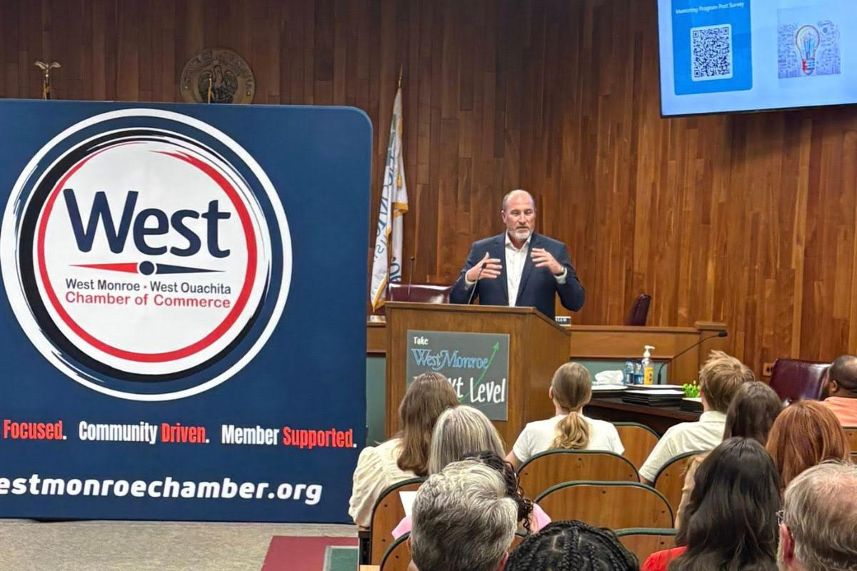A man speaks at a podium during a West Monroe-West Ouachita Chamber of Commerce event in a wood-paneled room, with seated attendees listening.