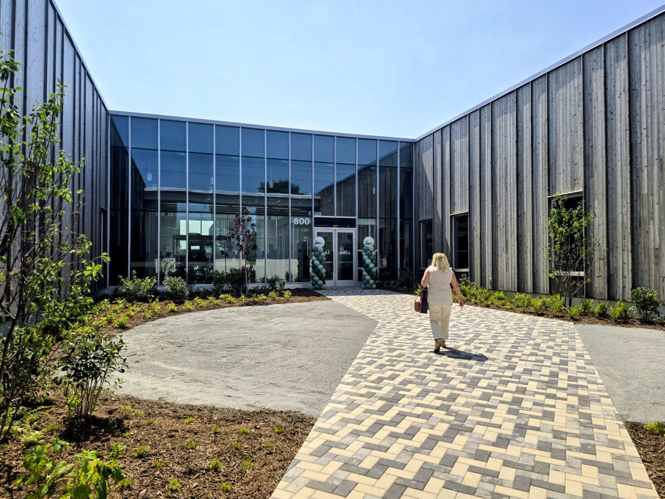 A person walks toward the glass entrance of a modern building with wood-paneled walls and a patterned brick pathway on a sunny day.