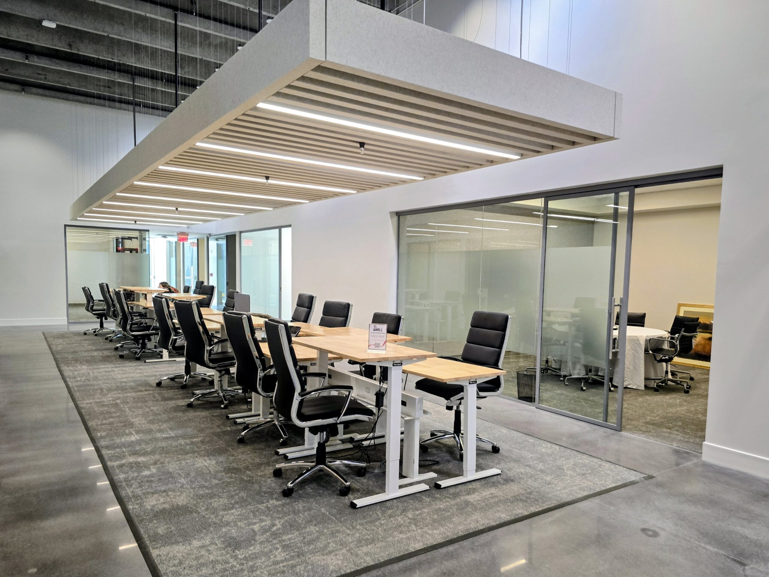 Modern open office space with long shared desks, black chairs, a large overhead light fixture, glass-walled meeting room, and polished concrete floors.