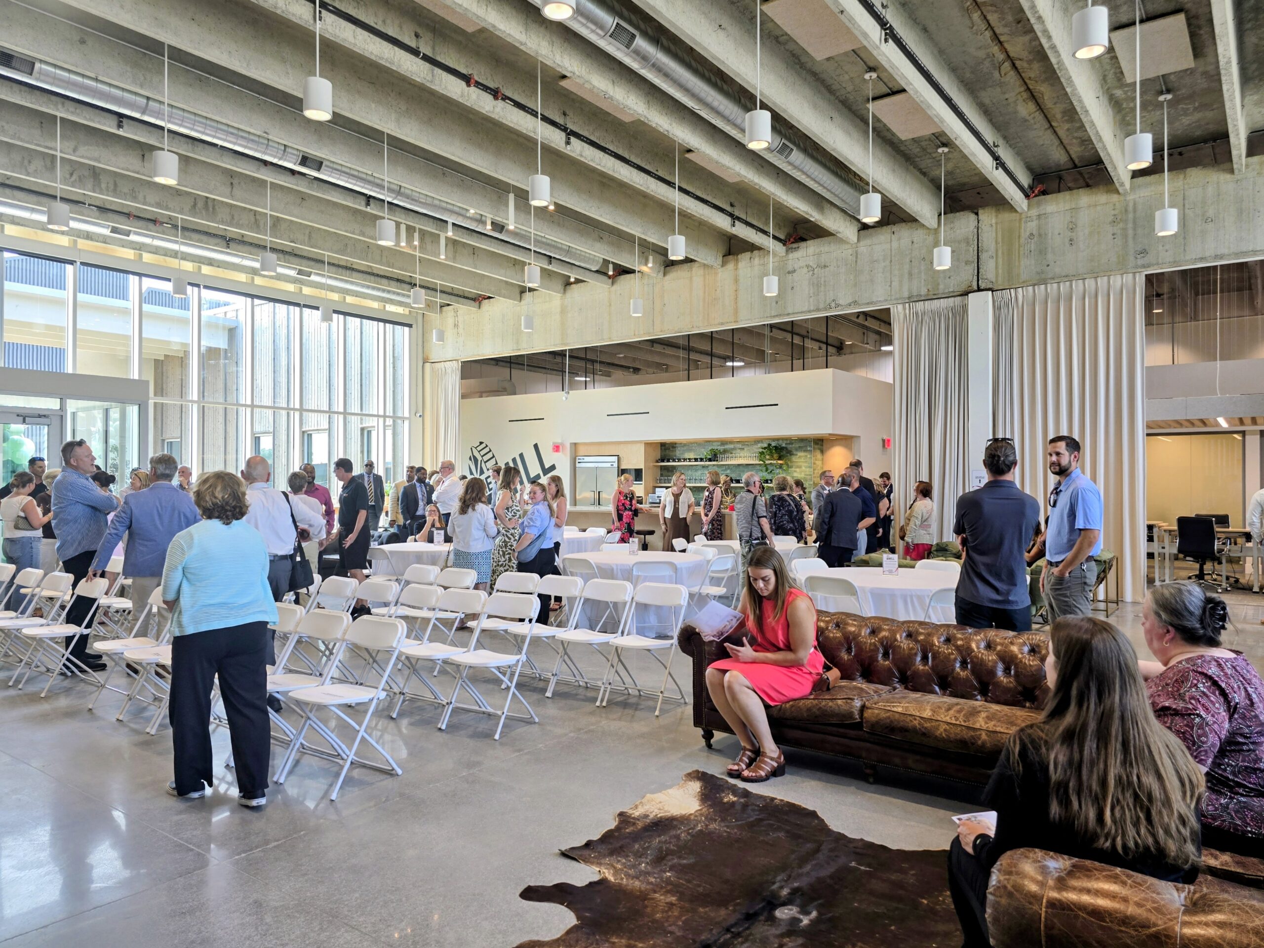 People gather in a modern, open room with folding chairs, a leather sofa, and exposed ceilings. Some are seated, while others stand and converse. Natural light fills the space.