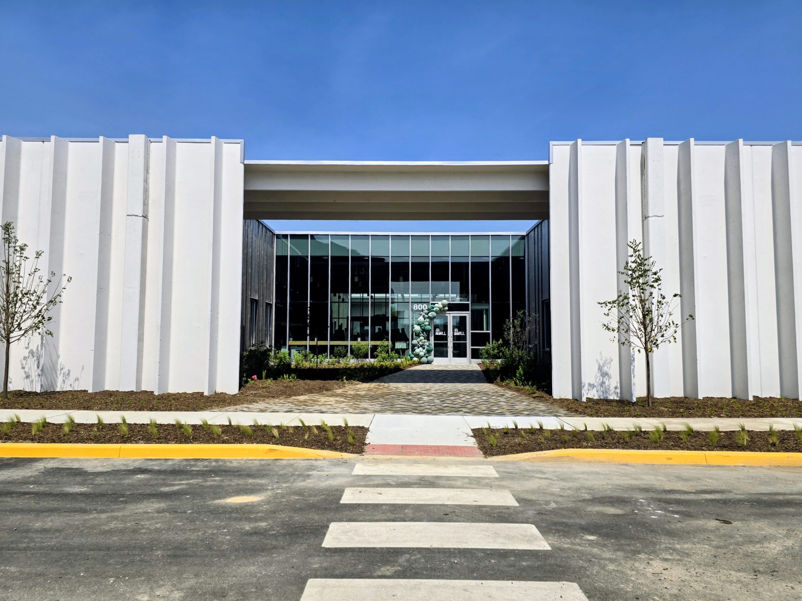 Modern building with tall white walls, large glass entrance, and newly landscaped grounds, viewed from across a street with a crosswalk under a clear blue sky.