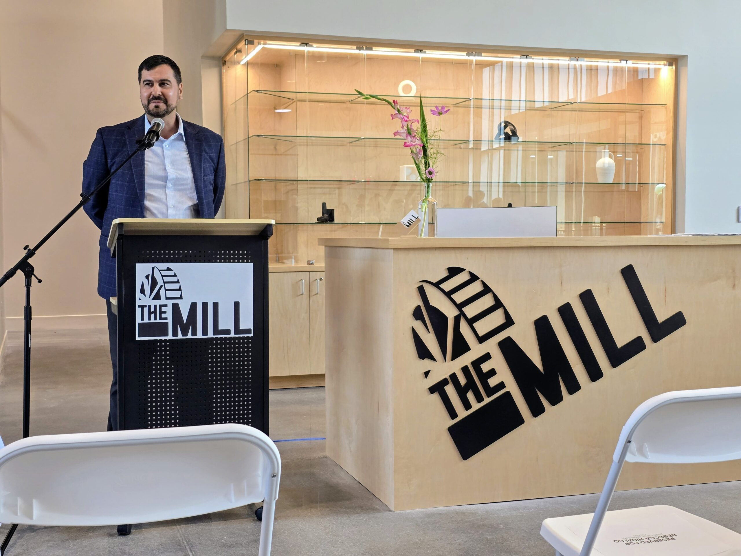 A man in a suit stands at a podium labeled "The Mill" next to a modern wooden reception desk with the same logo, in a bright room with chairs and shelves.
