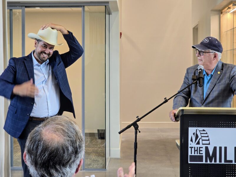 A man in a cowboy hat smiles while standing next to a man at a podium labeled "The Mill" during an indoor event.