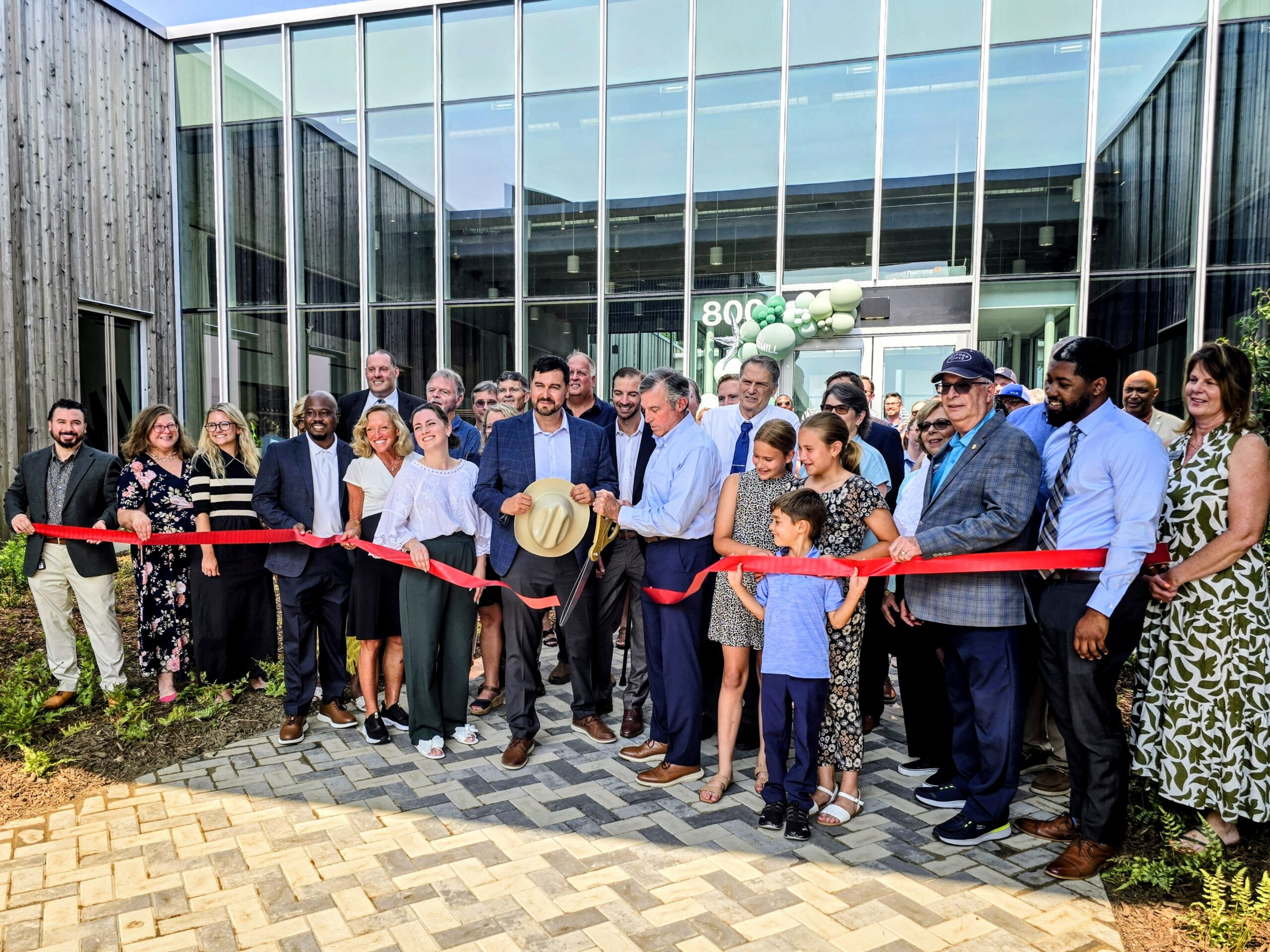 A group of people stands outside a modern building as a man cuts a red ribbon, celebrating a grand opening event.