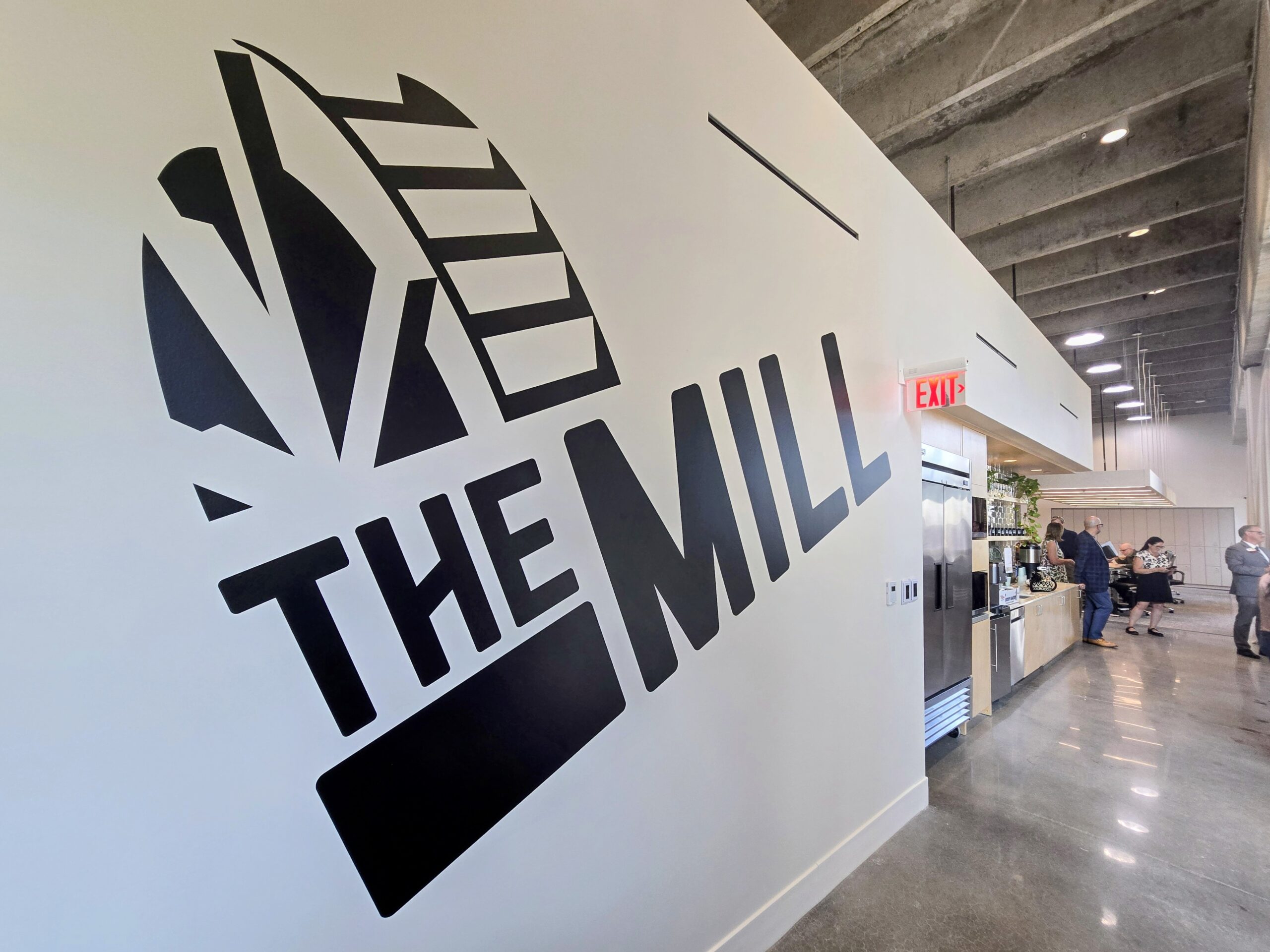 Large black "THE MILL" mural on a white wall inside a modern, industrial-style space with exposed ceiling and people gathered near a coffee area.