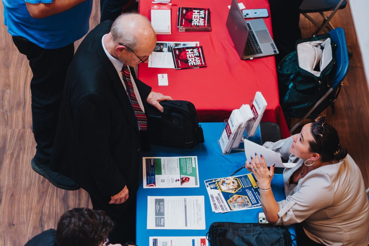 A man in a suit talks to a woman seated at a table covered with brochures, magazines, and a laptop at an indoor event.