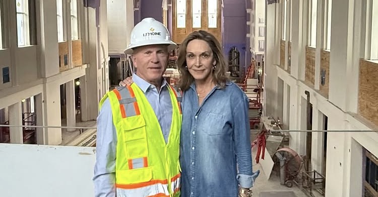 Two people stand side by side in a large building under construction; one wears a safety vest and hard hat, the other is in casual clothing.