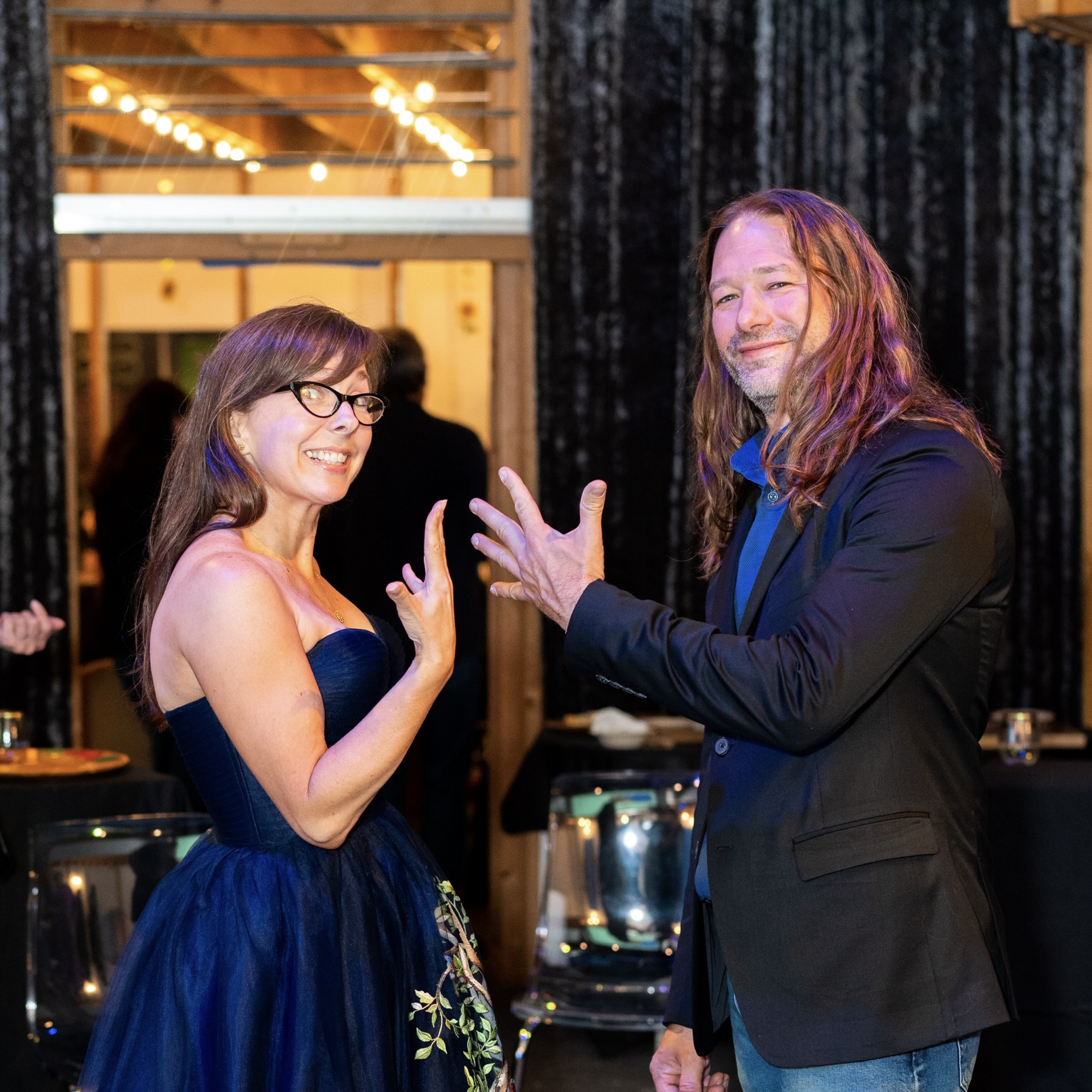A woman in a blue dress and a man in a dark jacket smile and gesture with their hands at an indoor event with string lights in the background.
