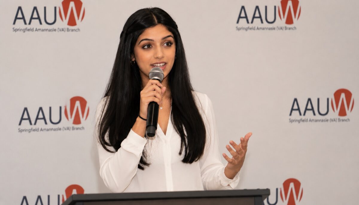 A woman in a white blouse speaks into a microphone at a podium, standing in front of a backdrop with AAUW Springfield Annasaise (VA) Branch logos.