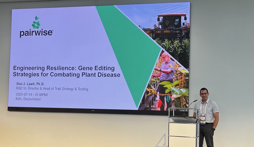 A man stands at a podium next to a large screen displaying a presentation titled "Engineering Resilience: Gene Editing Strategies for Combating Plant Disease" at a conference.
