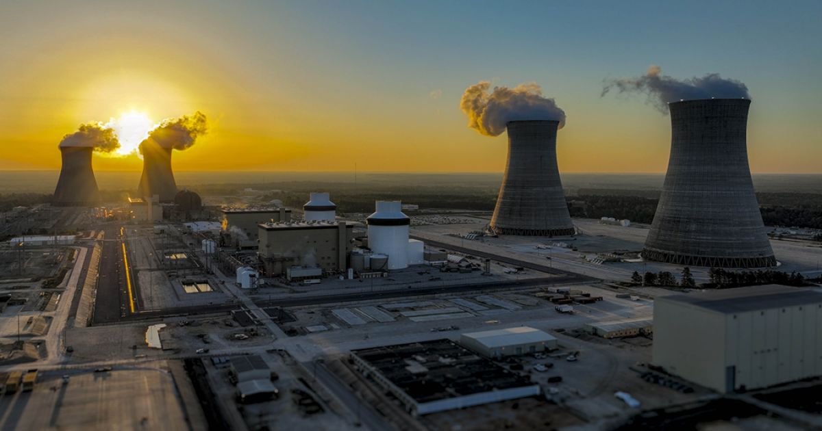 A nuclear power plant at sunset with cooling towers emitting steam, surrounded by industrial buildings and an expansive outdoor area.