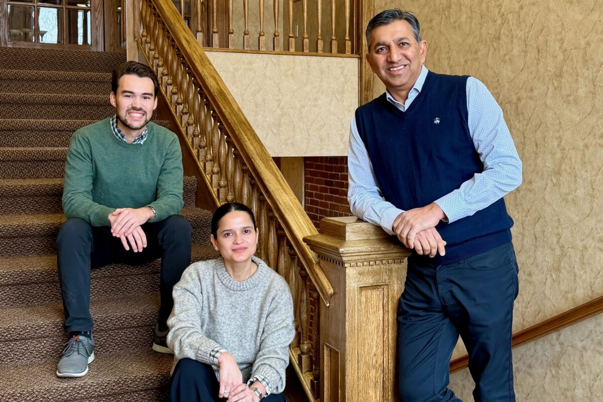 Three people pose on a carpeted staircase with a wooden bannister; two are sitting on the stairs and one is standing at the bottom, smiling at the camera.