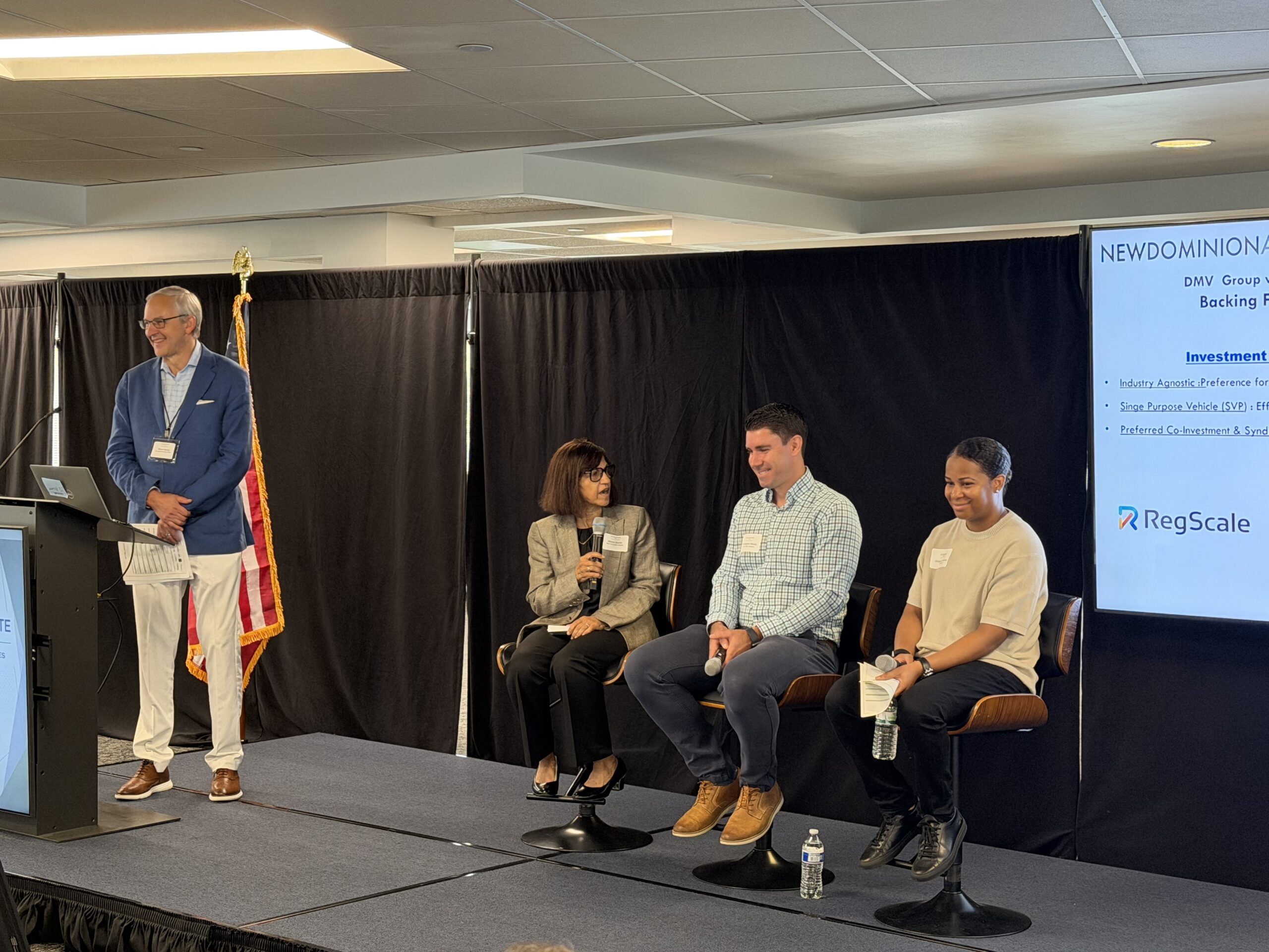 Four people participate in a panel discussion on a stage. Three are seated, one is standing at a podium. A screen displays event and sponsor information. An American flag is in the background.