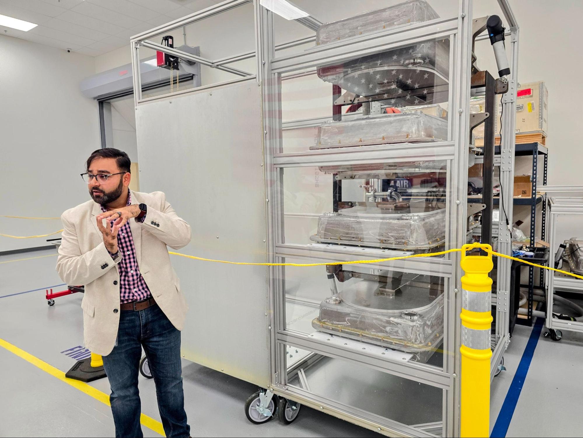 A man in a beige jacket gestures while standing next to a large metal rack holding covered trays in a laboratory setting with safety barriers.