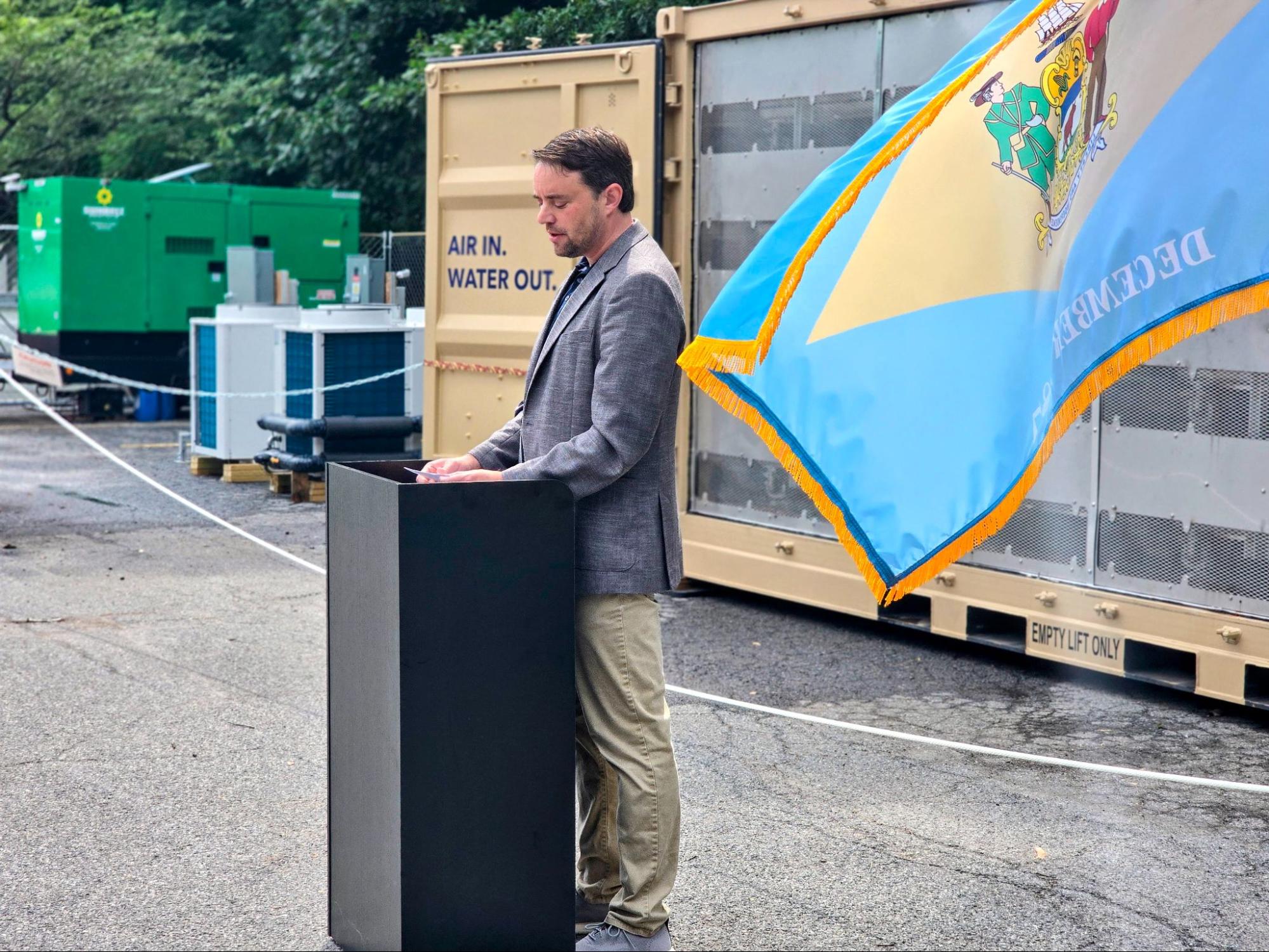 A man stands at a podium outdoors near containers labeled "AIR IN. WATER OUT." and a Delaware state flag, with equipment and trees in the background.