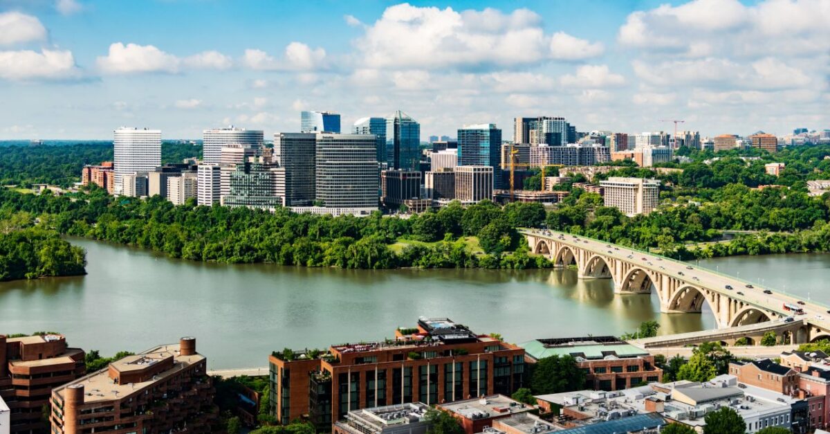 A city skyline with modern buildings, a river, a large bridge, and green trees under a partly cloudy sky.