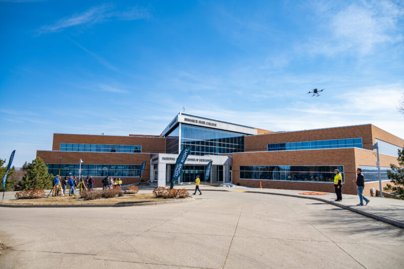 A modern brick building with large windows under a blue sky, people walking outside, and a drone flying above the entrance.