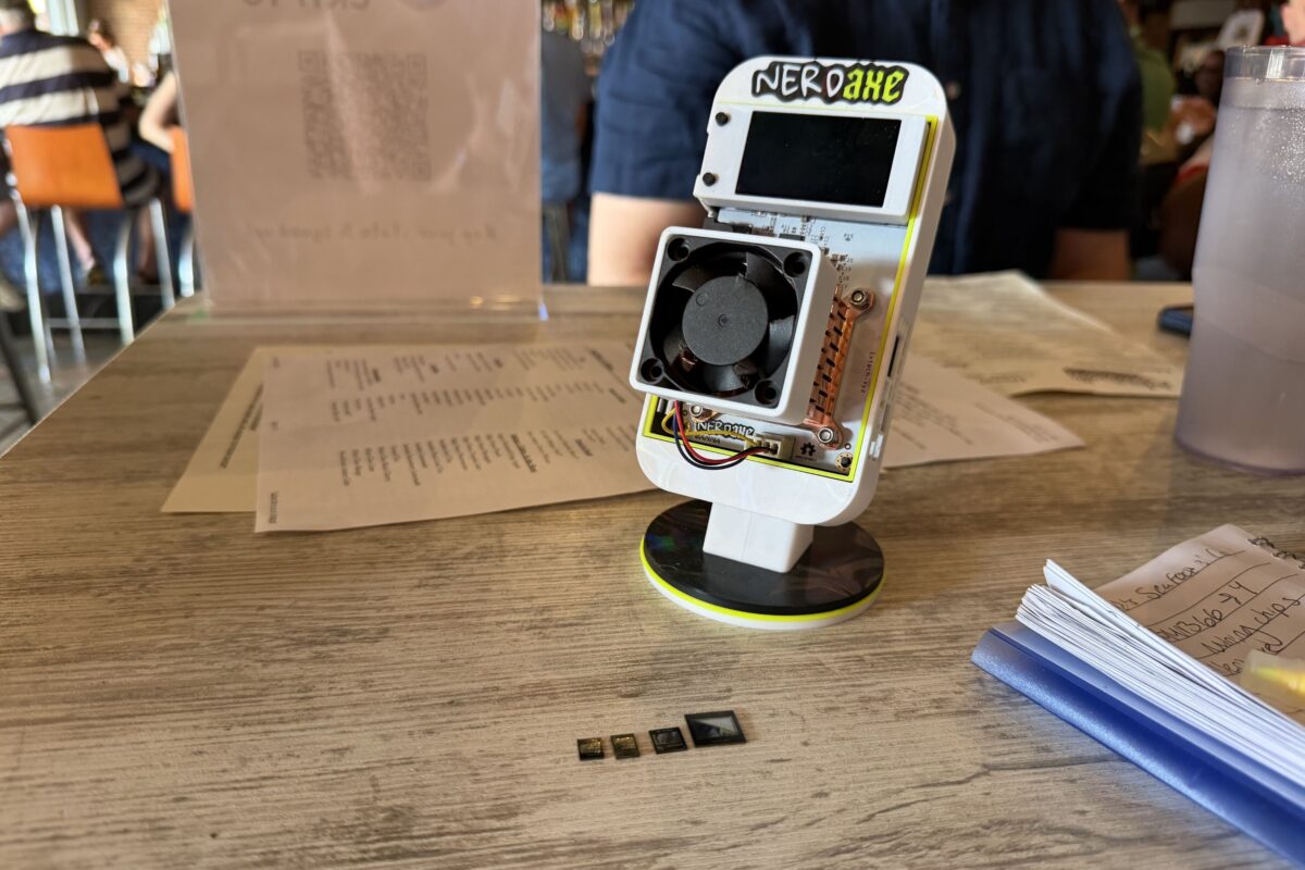 A small electronic device labeled "NERDahE" with a fan is displayed on a table, along with three tiny computer chips and a stack of papers in the background.