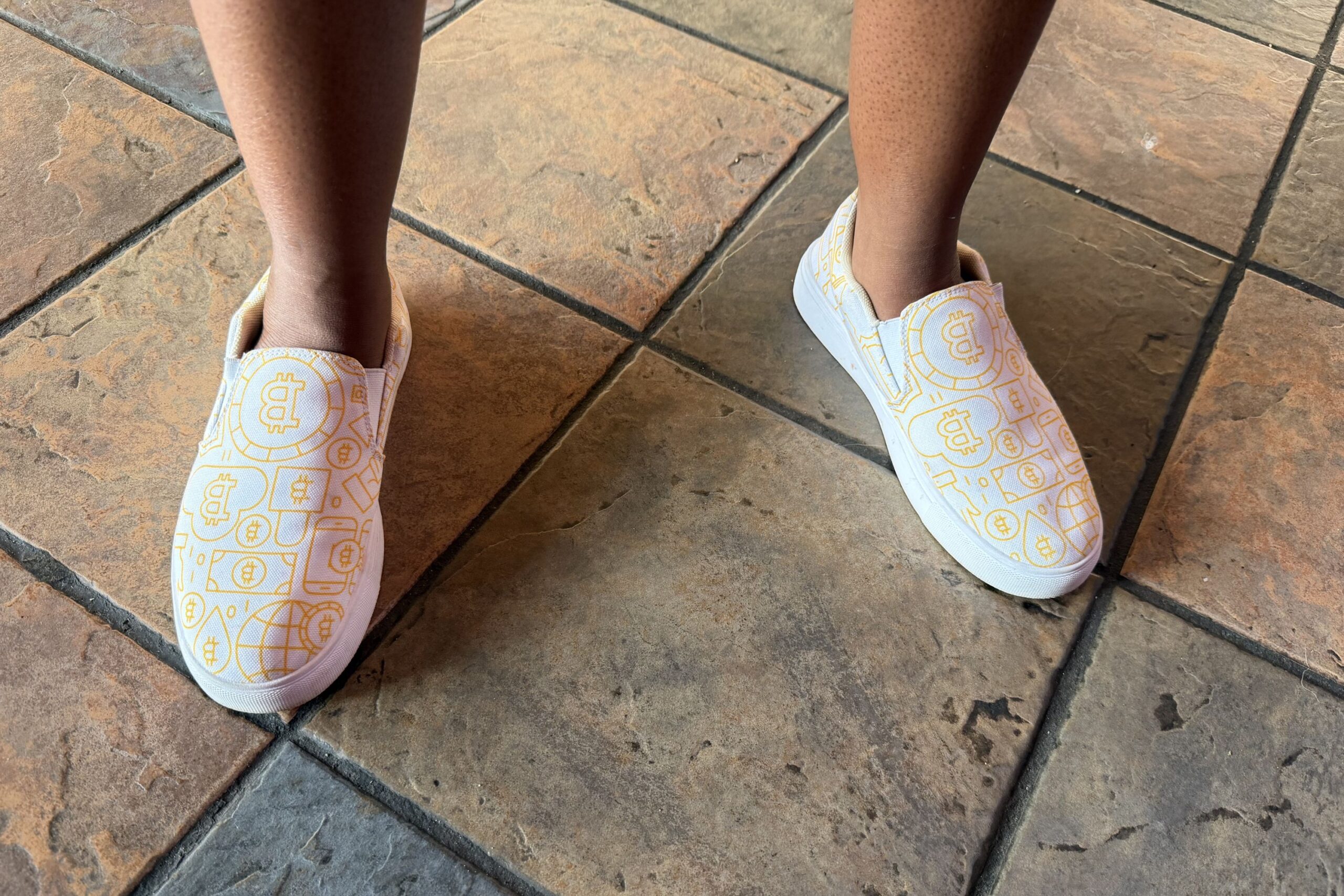 Person wearing white slip-on shoes with yellow geometric and currency symbol patterns, standing on a tiled floor.