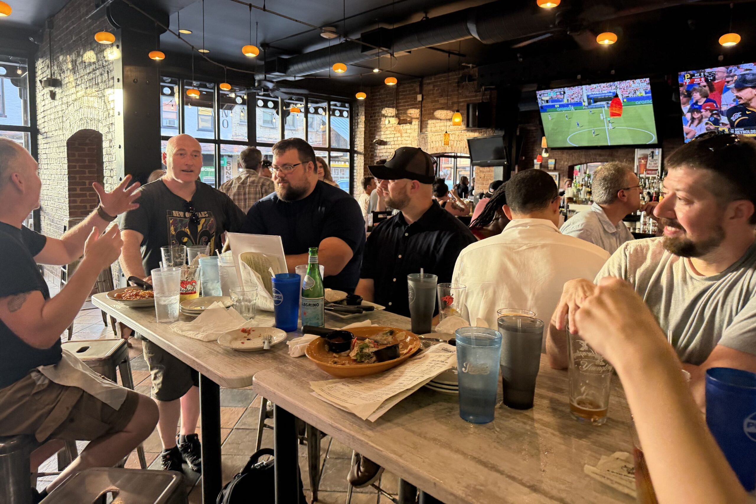 A group of people sit around a table with food and drinks in a casual restaurant or bar; several TV screens are visible in the background.