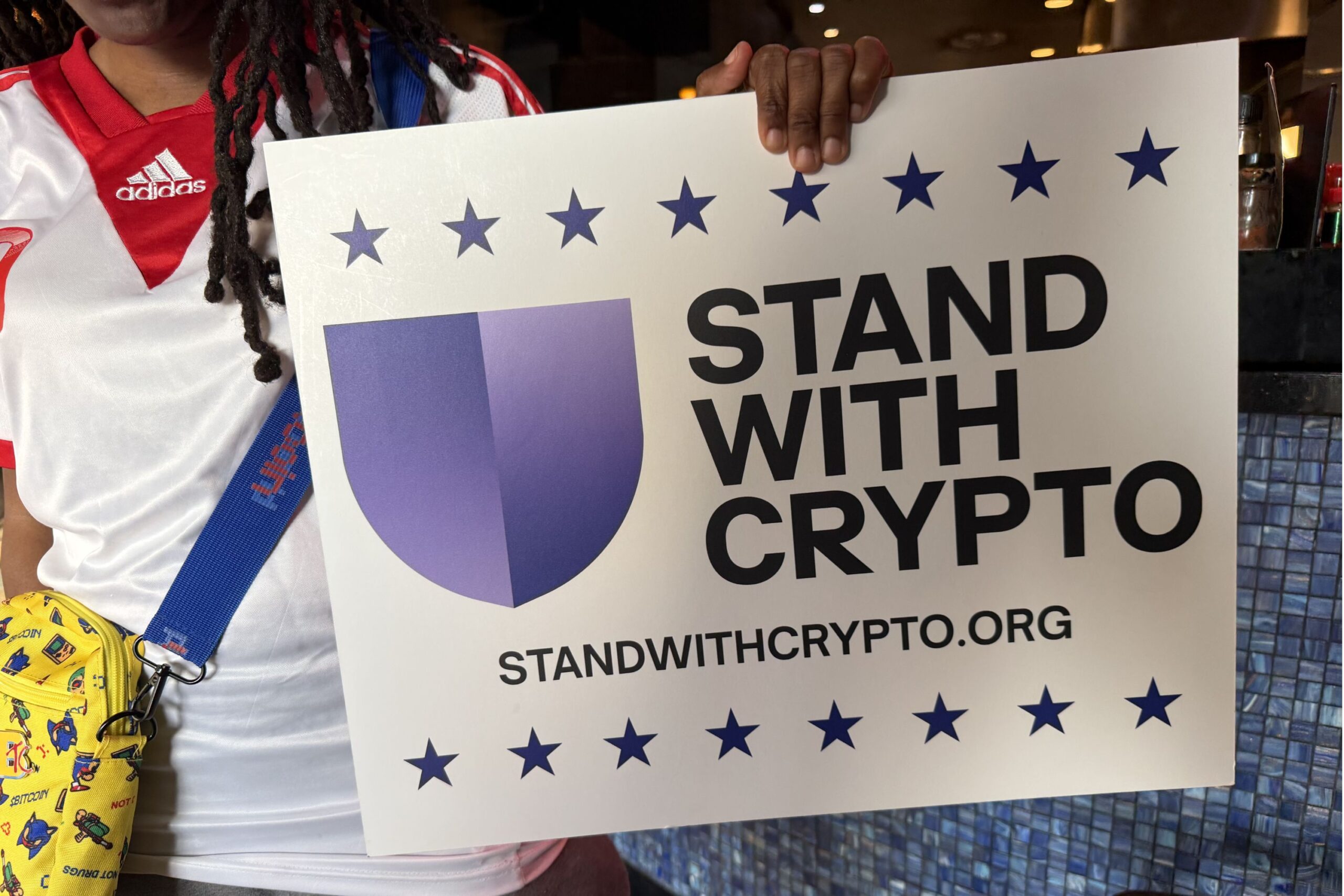 Person holding a sign that reads "STAND WITH CRYPTO" with a shield logo and website standwithcrypto.org, surrounded by blue stars.