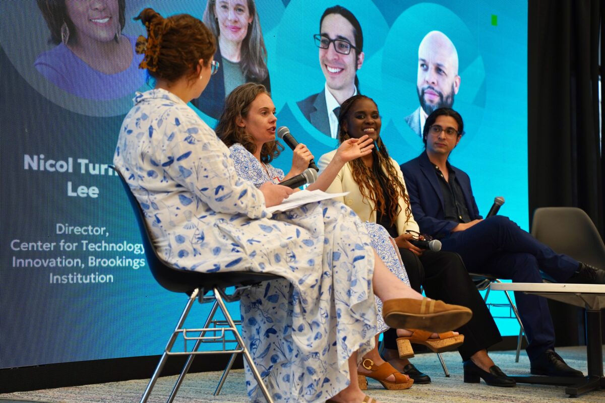 Four people sit on a stage panel discussing, with a blue screen behind them displaying headshots and information about one of the speakers. One woman speaks into a microphone.