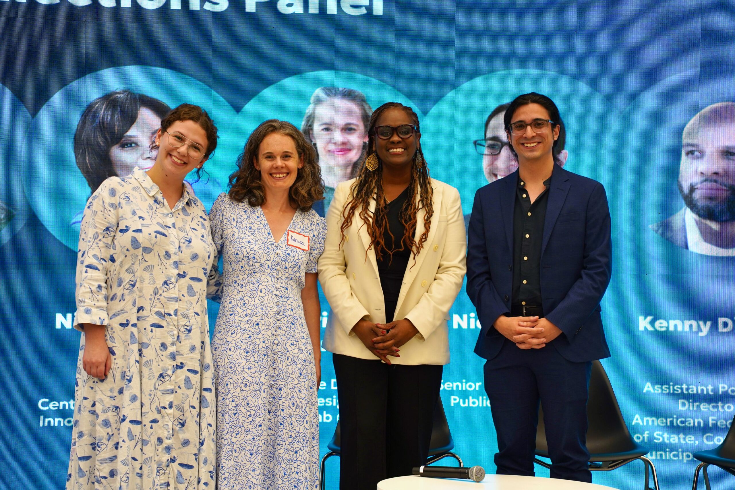 Four people stand and smile in front of a blue screen displaying headshots and text about a panel discussion. Two women wear patterned dresses, one woman wears a white suit, and one man wears a dark suit.