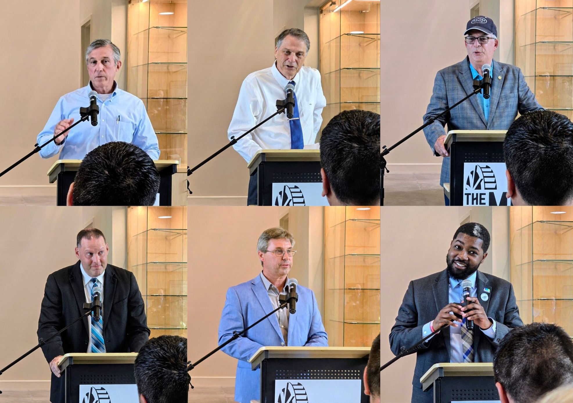 Six men, each at a podium with microphones, speak to an audience in a professional indoor setting.