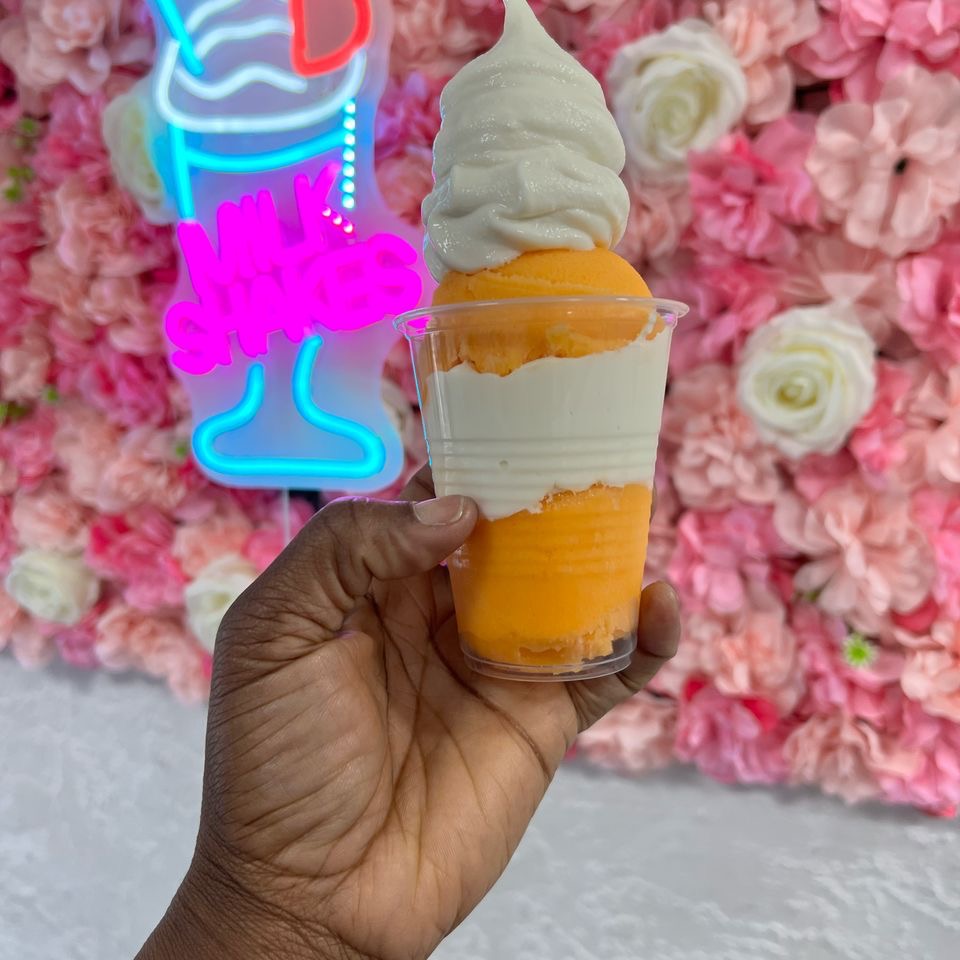 A hand holds a cup with orange and white soft serve ice cream in front of a pink floral wall and a neon milkshakes sign.
