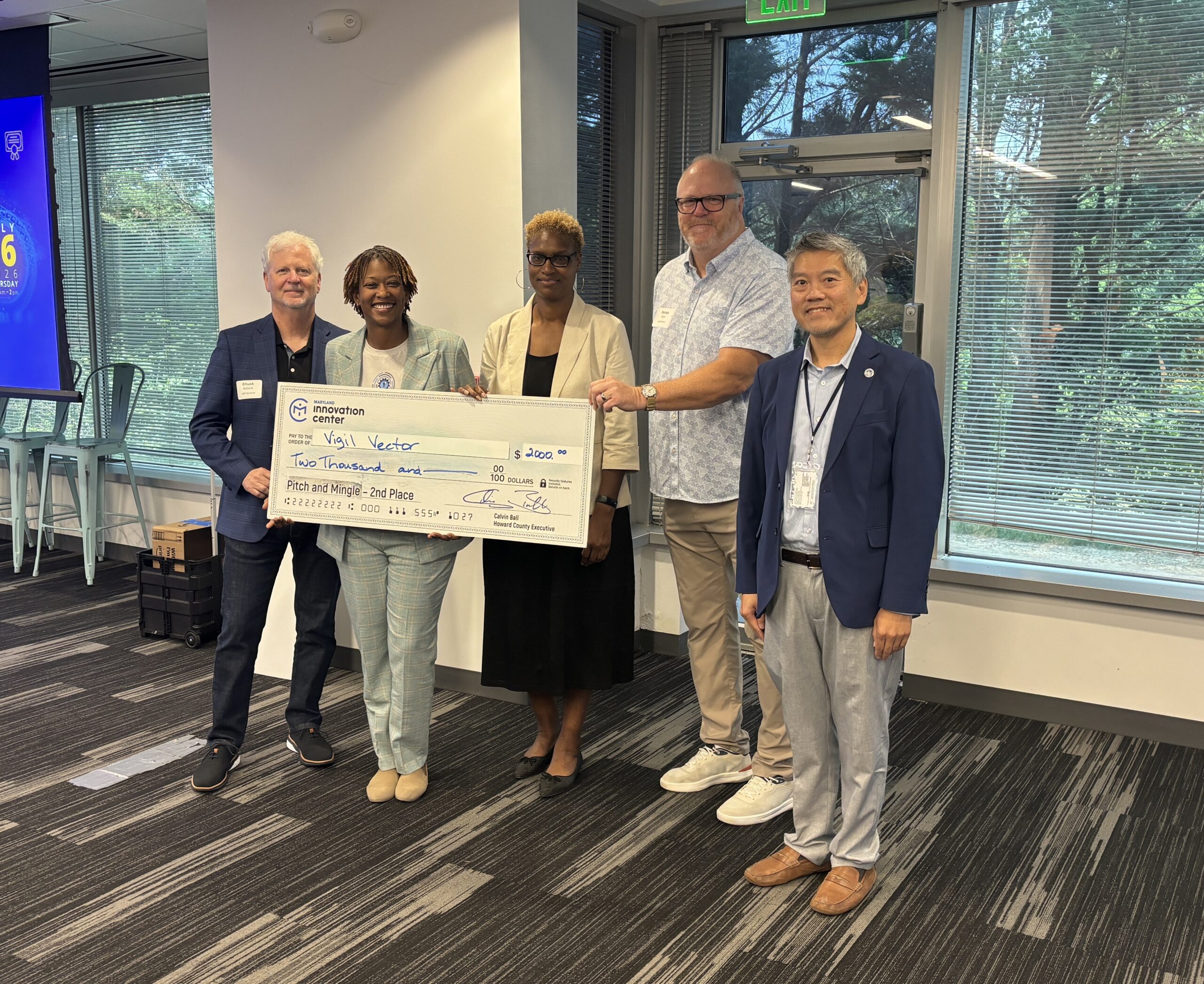 Five people stand indoors holding a large check for $250,000 made out to the organization "Vigil Vector" at a formal event.