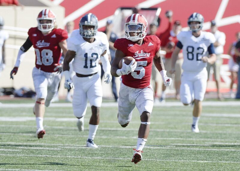 Indiana football player in red runs with the ball down the field as multiple opponents in white uniforms chase him during a game.