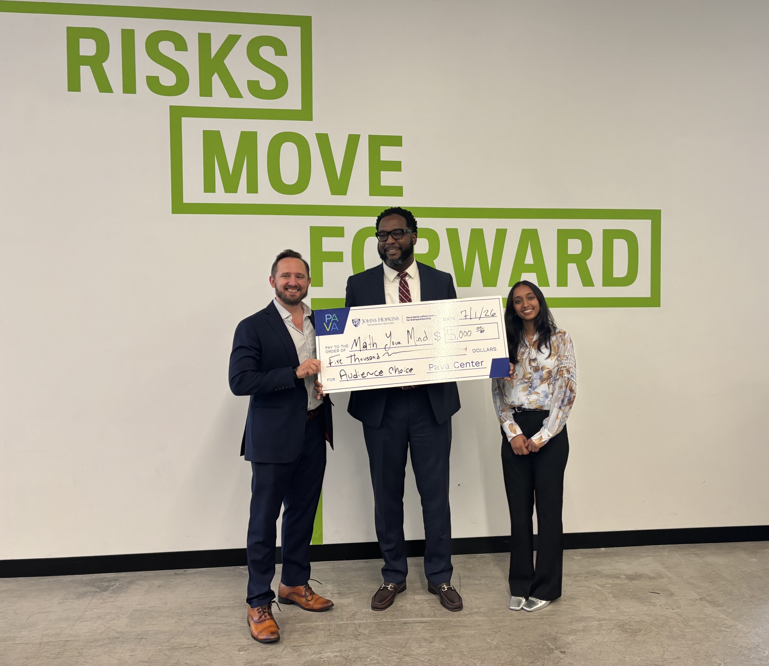 Three people stand indoors holding a large check for $5,000, with "Risks Move Forward" on the wall behind them.